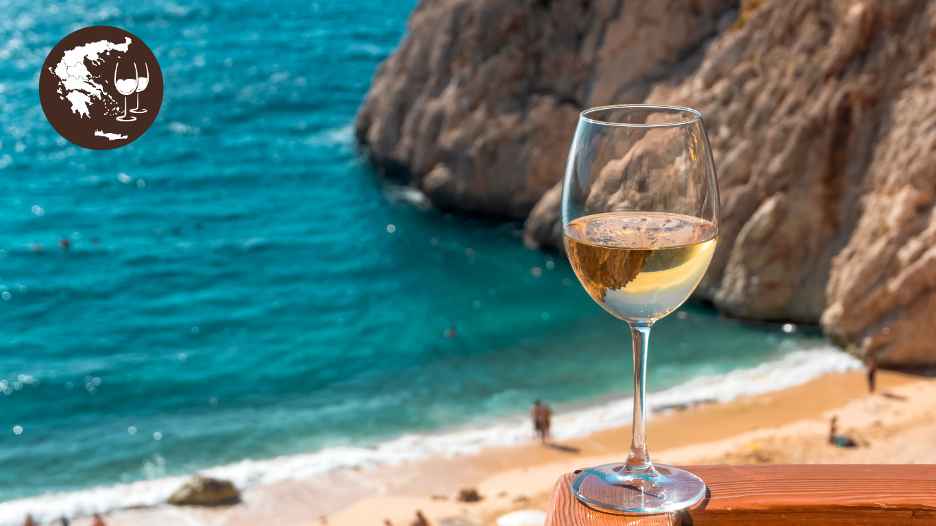 Weißwein im Glas vor einem Strand mit türkisfarbenem Wasser und Felsen im Hintergrund.