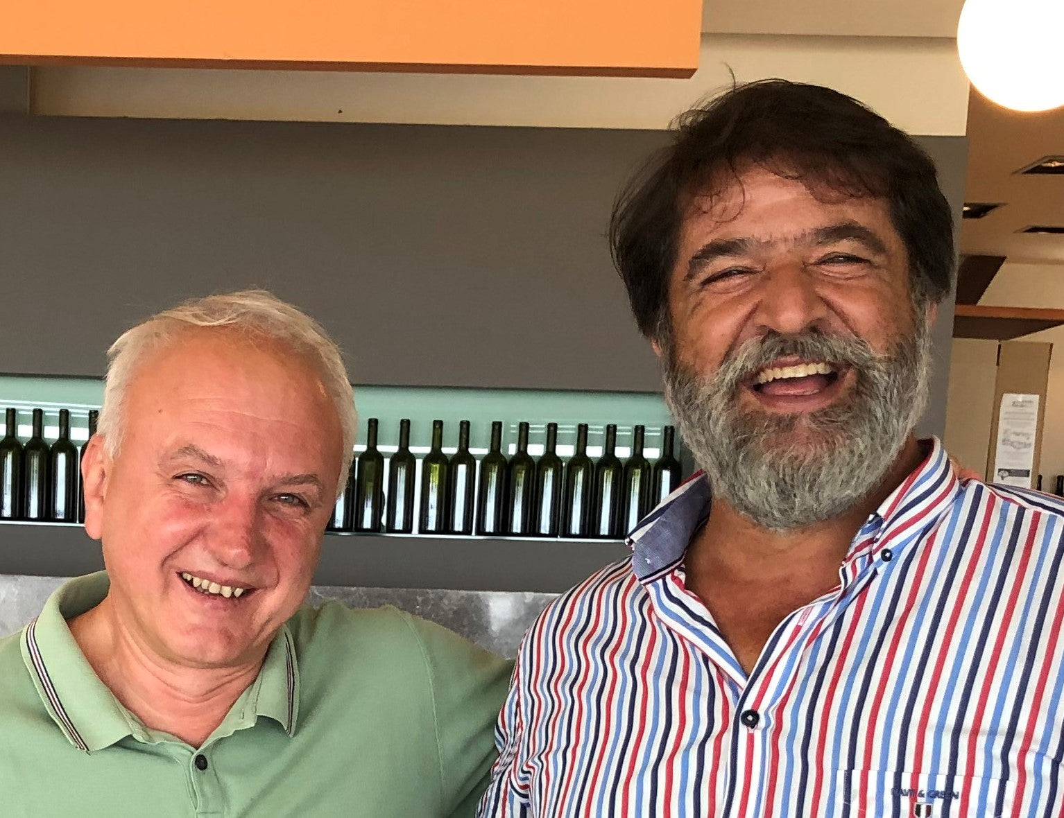 Zwei Männer lächeln in einem Restaurant mit einer Reihe von Flaschen im Hintergrund.