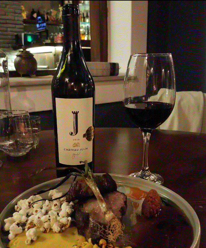 Chateau Julia Merlot 2023 Flasche mit Glas Rotwein und Gourmetgericht auf Holztisch