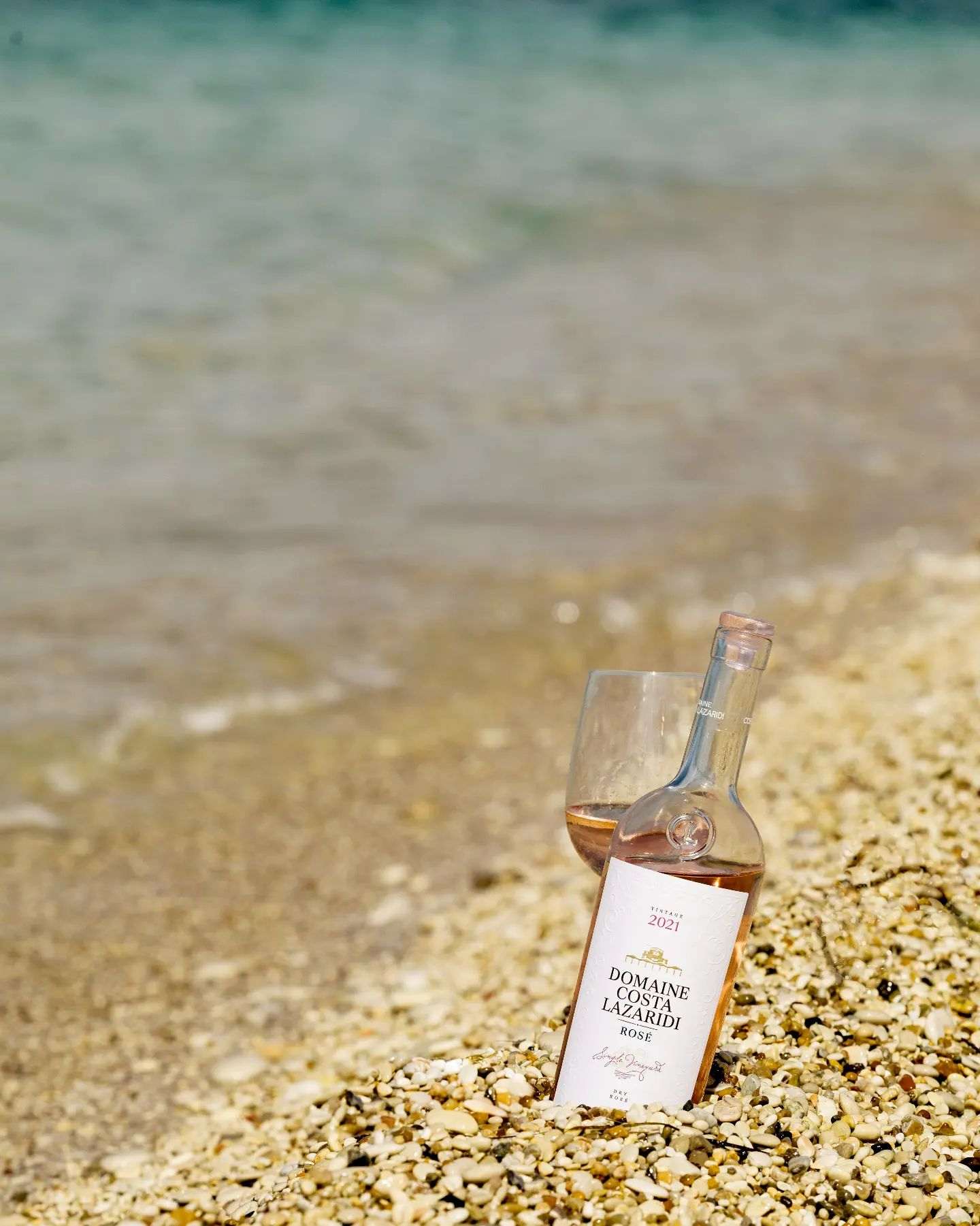 Flasche Domaine Costa Lazaridi Rosé 2024 mit Glas am Kieselstrand und Meer im Hintergrund