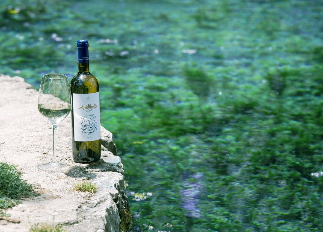 Flasche Amethystos White Weißwein und Weinglas auf Steinmauer mit grünem Wasser im Hintergrund