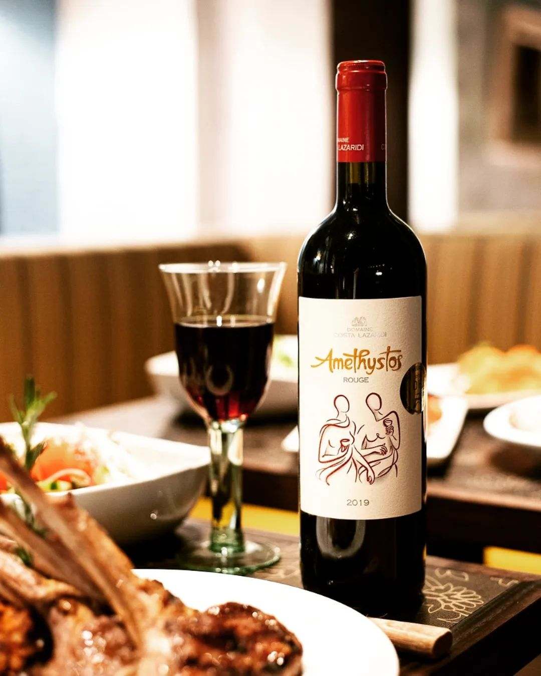 Amethystos Red Rotwein Flasche mit Glas und Lammgericht in Restaurantumgebung