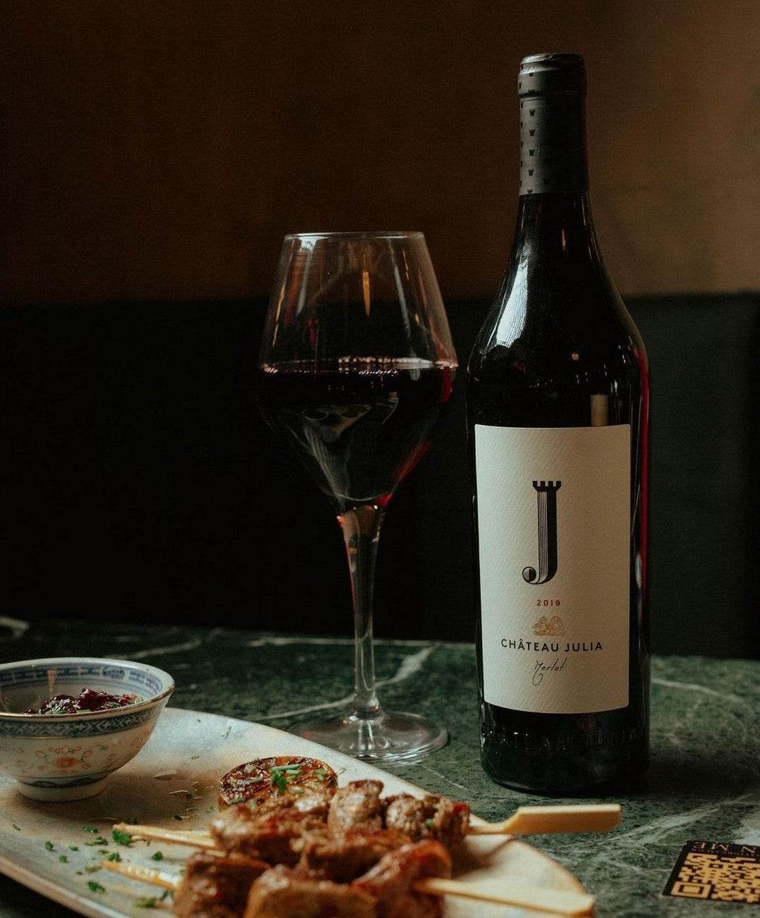 Chateau Julia Merlot 2023 Flasche und Glas mit dunklem Rotwein und Grillspießen auf Tisch