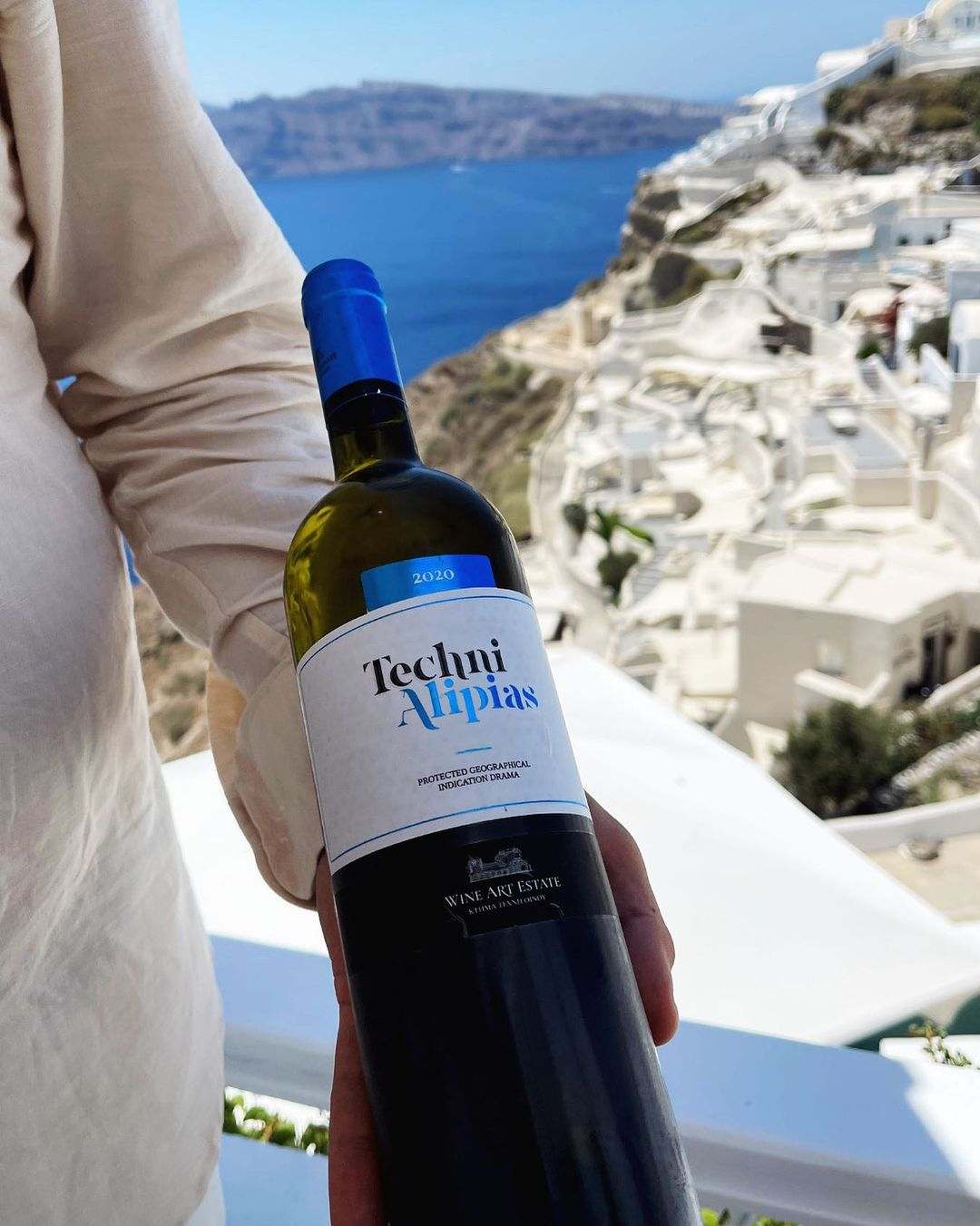 Person hält Techni White Weinflasche mit blauem Verschluss vor mediterraner Küstenlandschaft