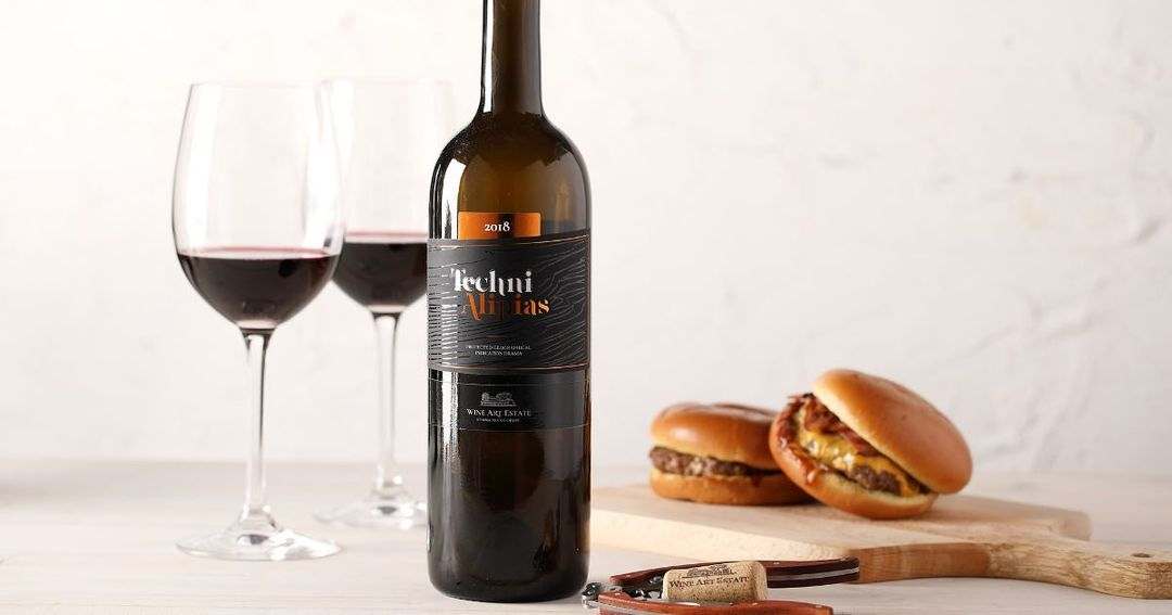 Flasche Techni Red Rotwein mit zwei Gläsern und Burgern auf hellem Tisch