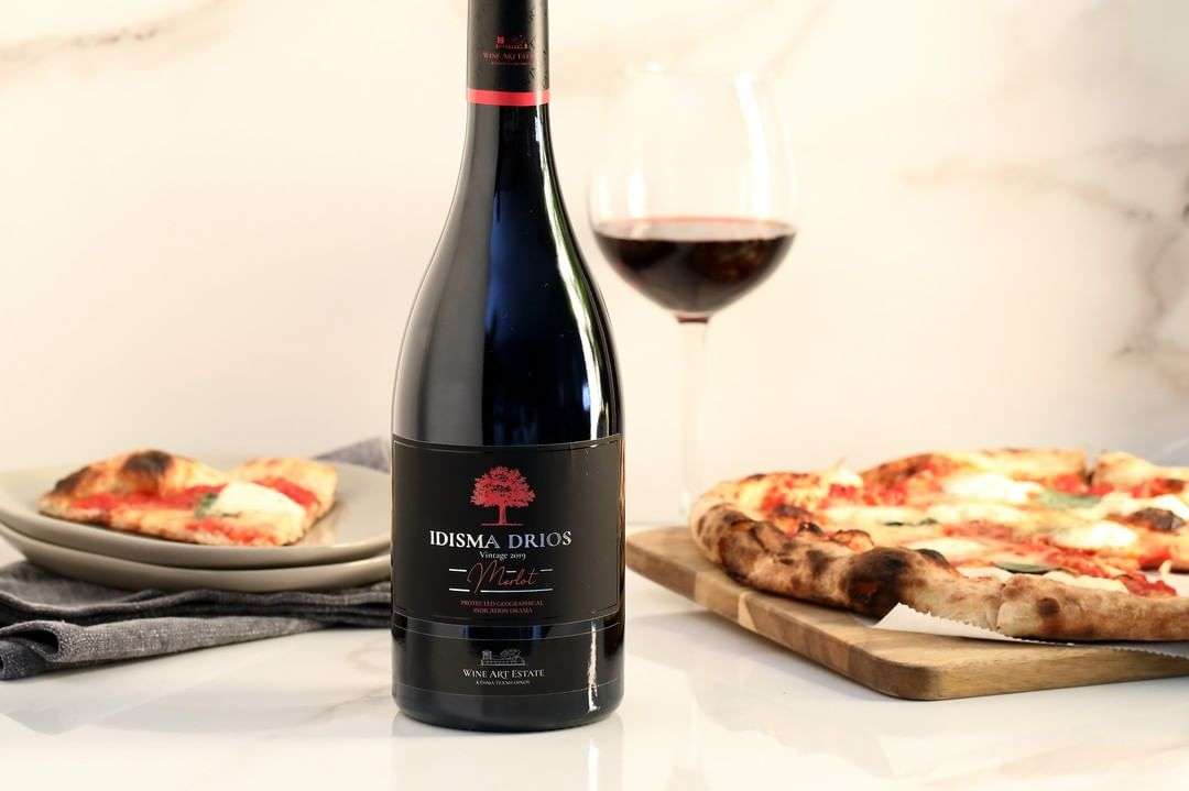 Flasche Idisma Drios Merlot 2021 mit Glas Rotwein und Pizza auf weißer Marmoroberfläche