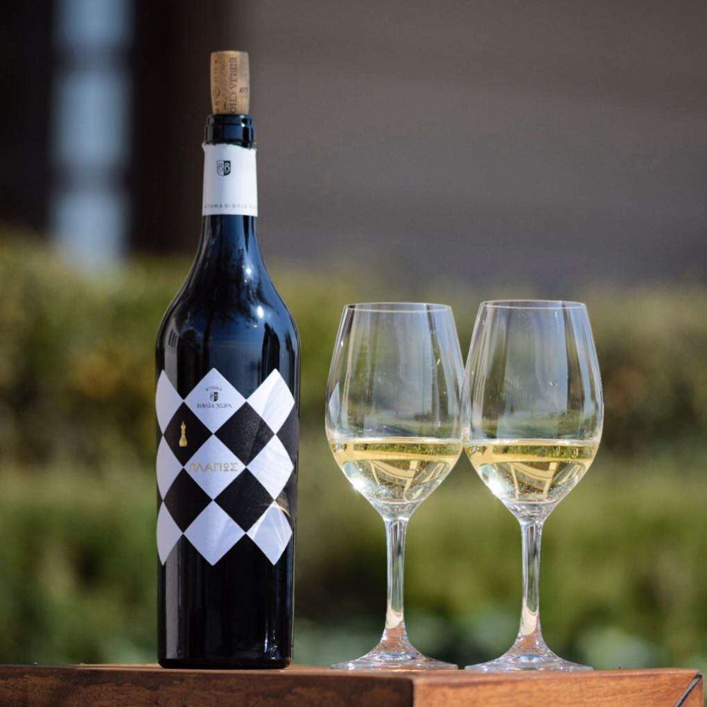 Biblia Chora Plagios White Chardonnay Weißwein 2022 mit zwei gefüllten Weingläsern auf Holzplatte im Freien