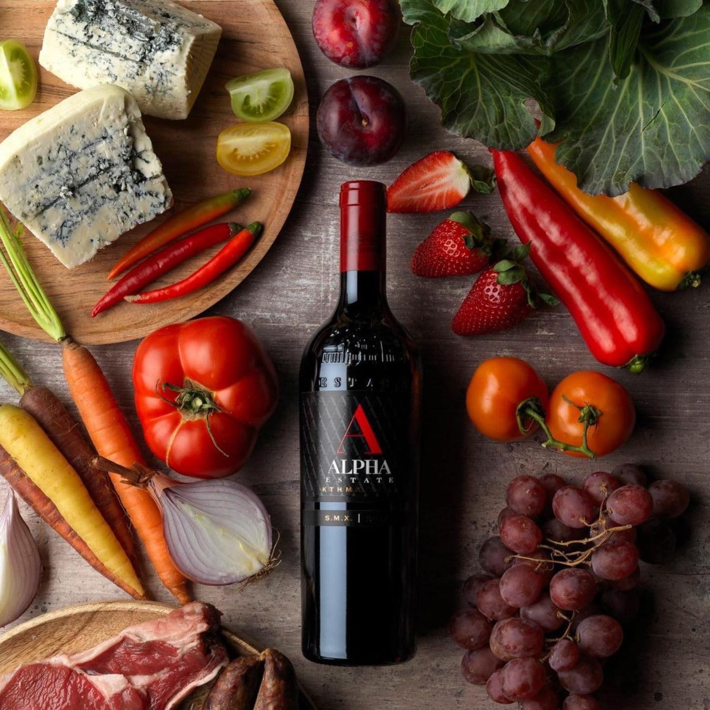 Flasche Alpha Estate SMX Rotwein umgeben von Käse, Gemüse, Obst und Fleisch auf Holzuntergrund