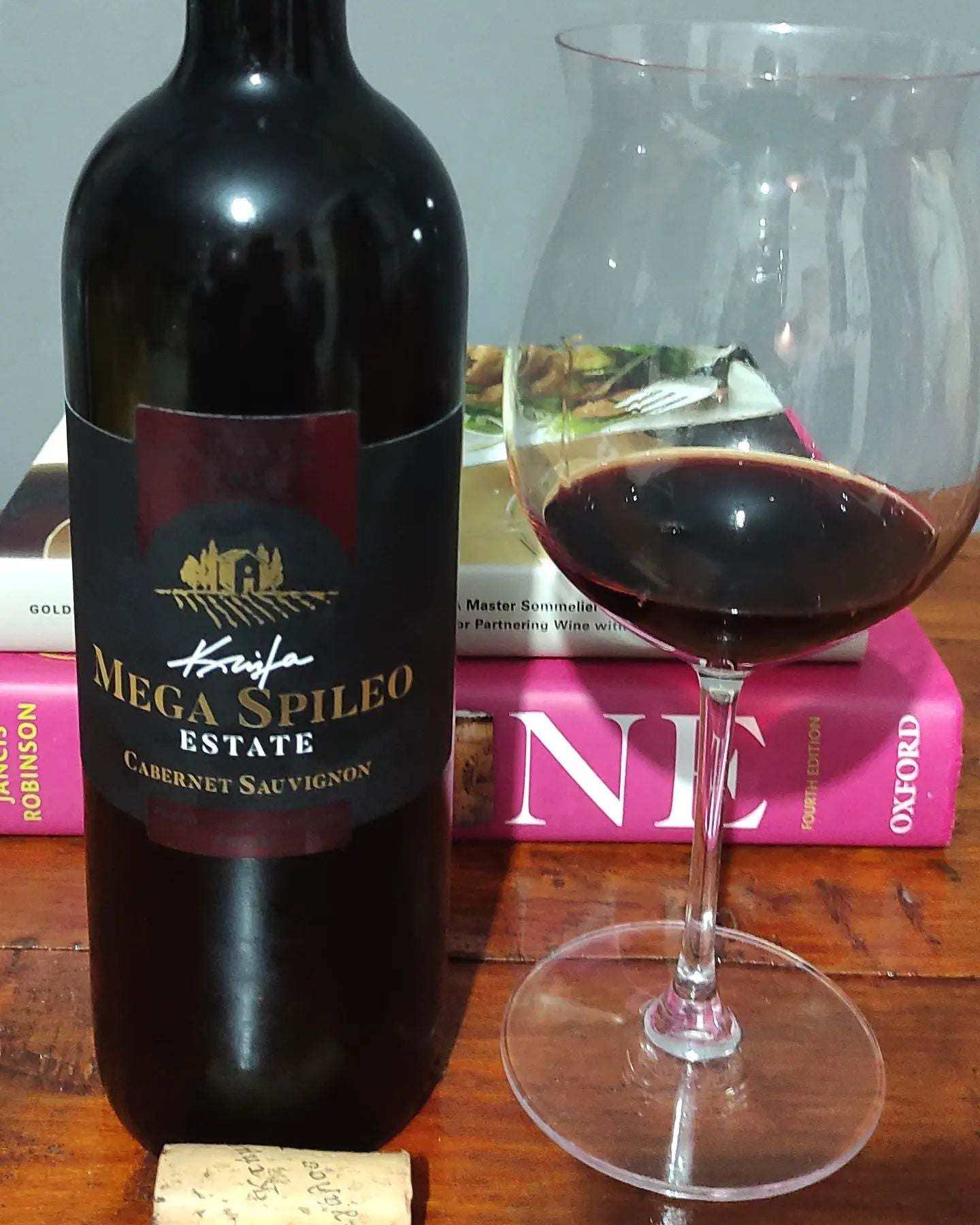 Flasche Mega Spileo Cabernet Sauvignon mit Glas tiefrotem Rotwein auf Holztisch vor Büchern