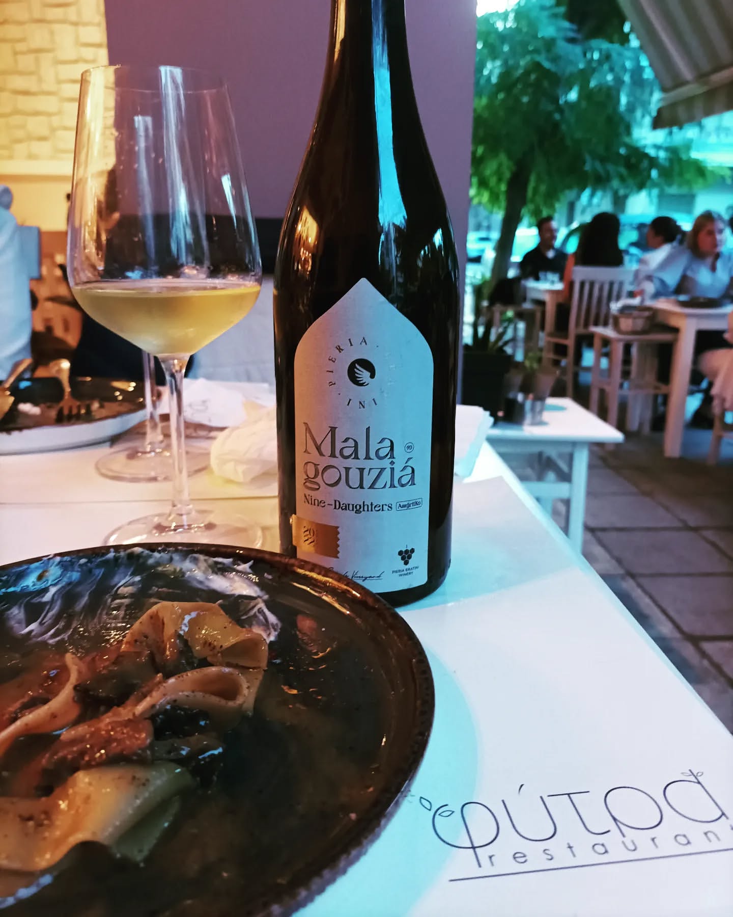 9 Daughters Malagousia von PIERIA ERATINI in einer eleganten Flasche, serviert mit einem Gericht auf einem Tisch.