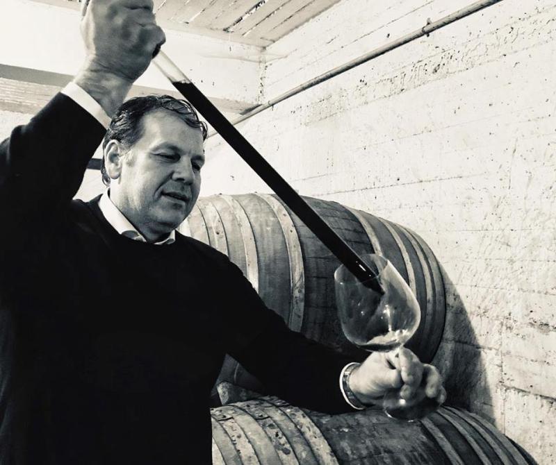 Mann mit einem Glas Wein, der mit einem Dekantierstab aus Metall Wein aus einem Fass einschenkt.