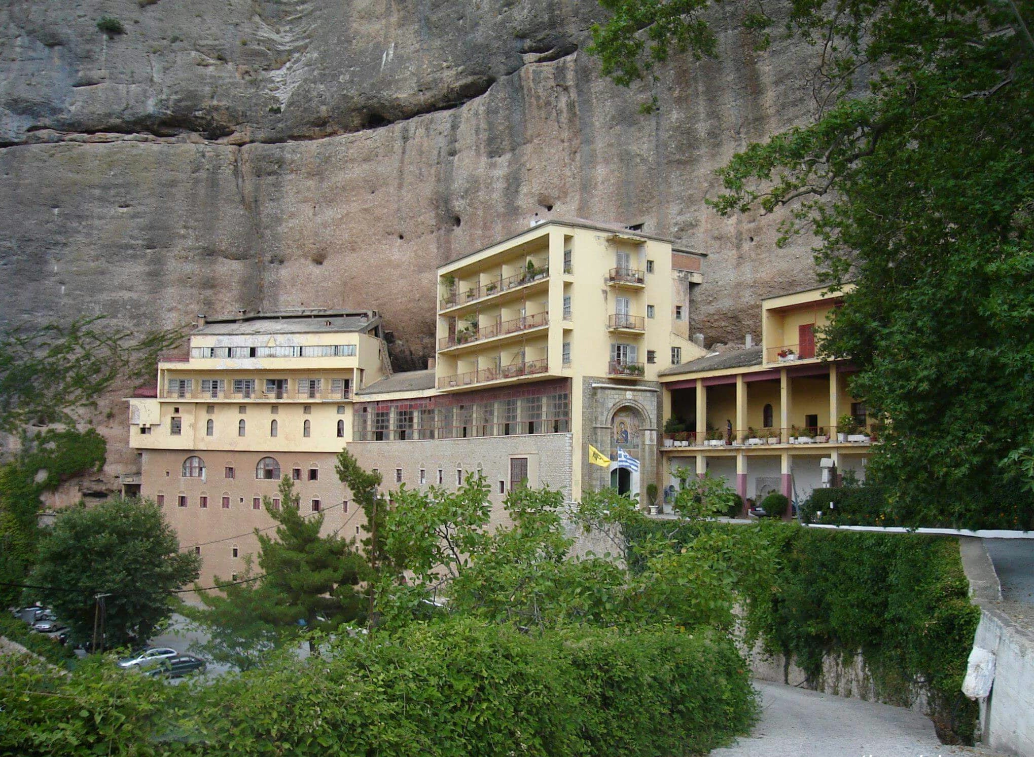 Klostergebäude in gelber und grauer Fassade, eingebettet in eine Felslandschaft mit Bäumen im Vordergrund.