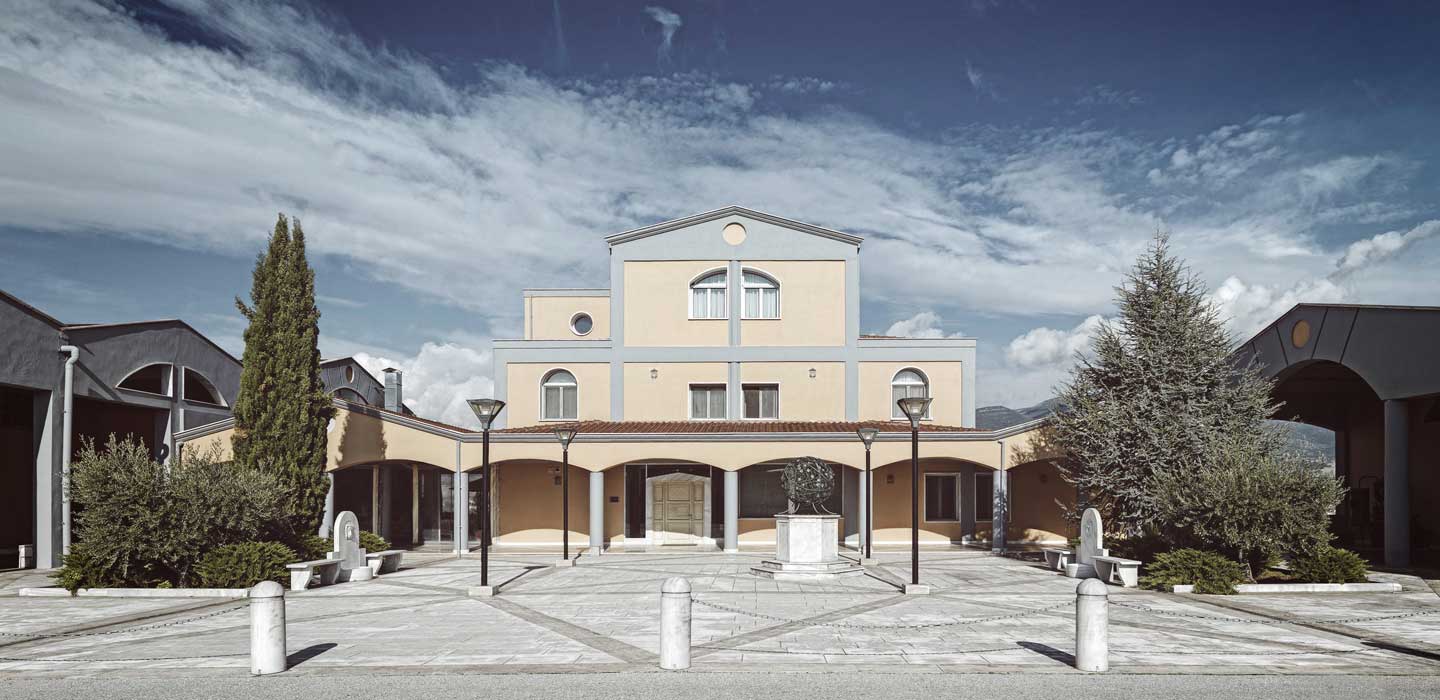 Gebäude mit gelber Fassade und großen Fenstern, umgeben von Bäumen und einer gepflasterten Fläche.