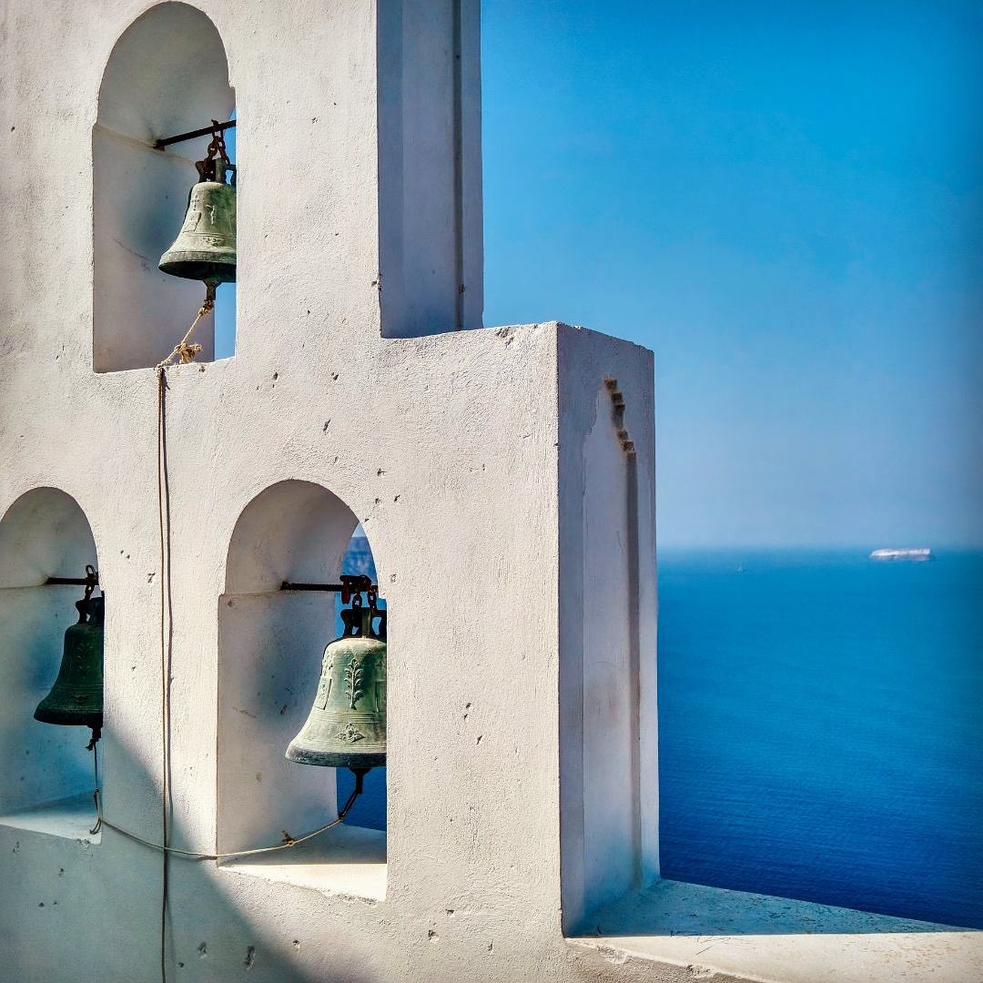 Drei grüne Glocken in einer weißen Kirchenarchitektur mit Blick auf das blaue Meer.