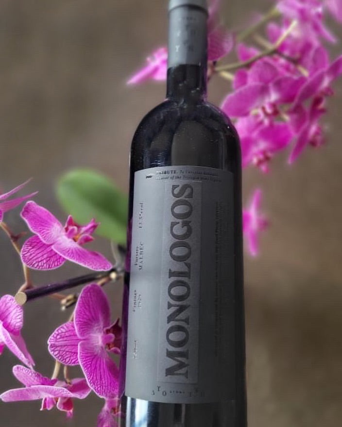 Monologos Malbec von Dyo Ipsi Estate in schwarzer Flasche mit Holzfass Ausbau und rosa Orchideen im Hintergrund