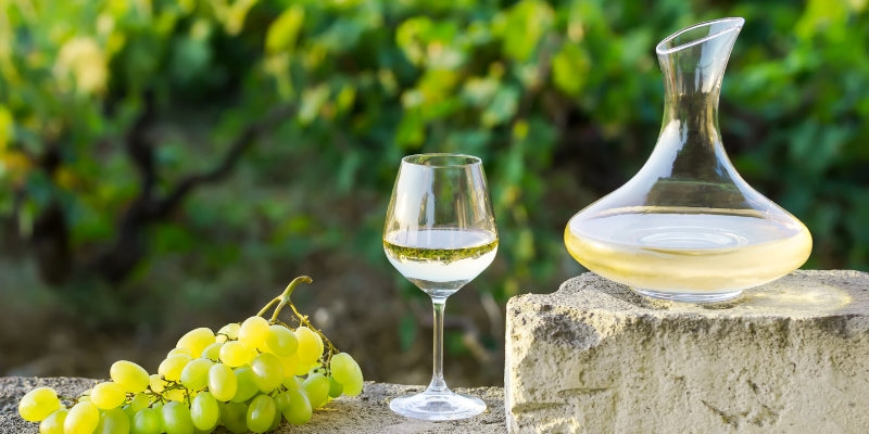 Weißwein in einer Glaskaraffe mit einem Weinglas und einer Traube auf einem Stein