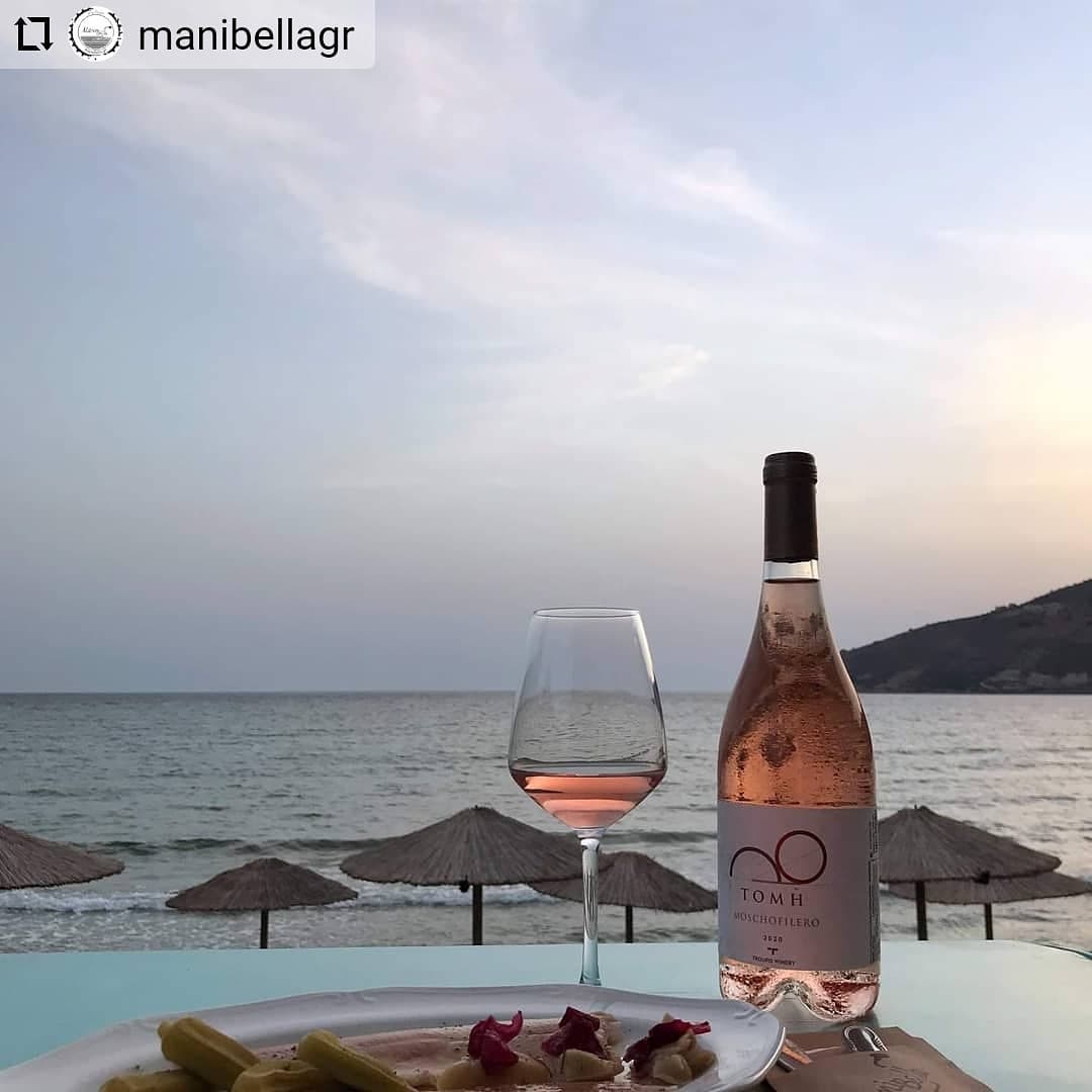 Köstlicher Sommerroséwein von Troupis Winery &quot;Tomi Rosé&quot; an paradiesischem Strand mit Blick aufs Meer. Ein Glas Roséwein und ein geschliffenes Kristallglas auf einem Steintisch umgeben von Sonnenschirmen. Die Flasche mit dem eleganten Design und der klaren Etikette steht im Fokus.